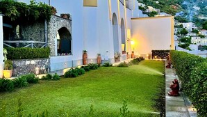 Um edifício branco com janelas em arco e uma parede de pedra, rodeado por vegetação e ao fundo, uma montanha.