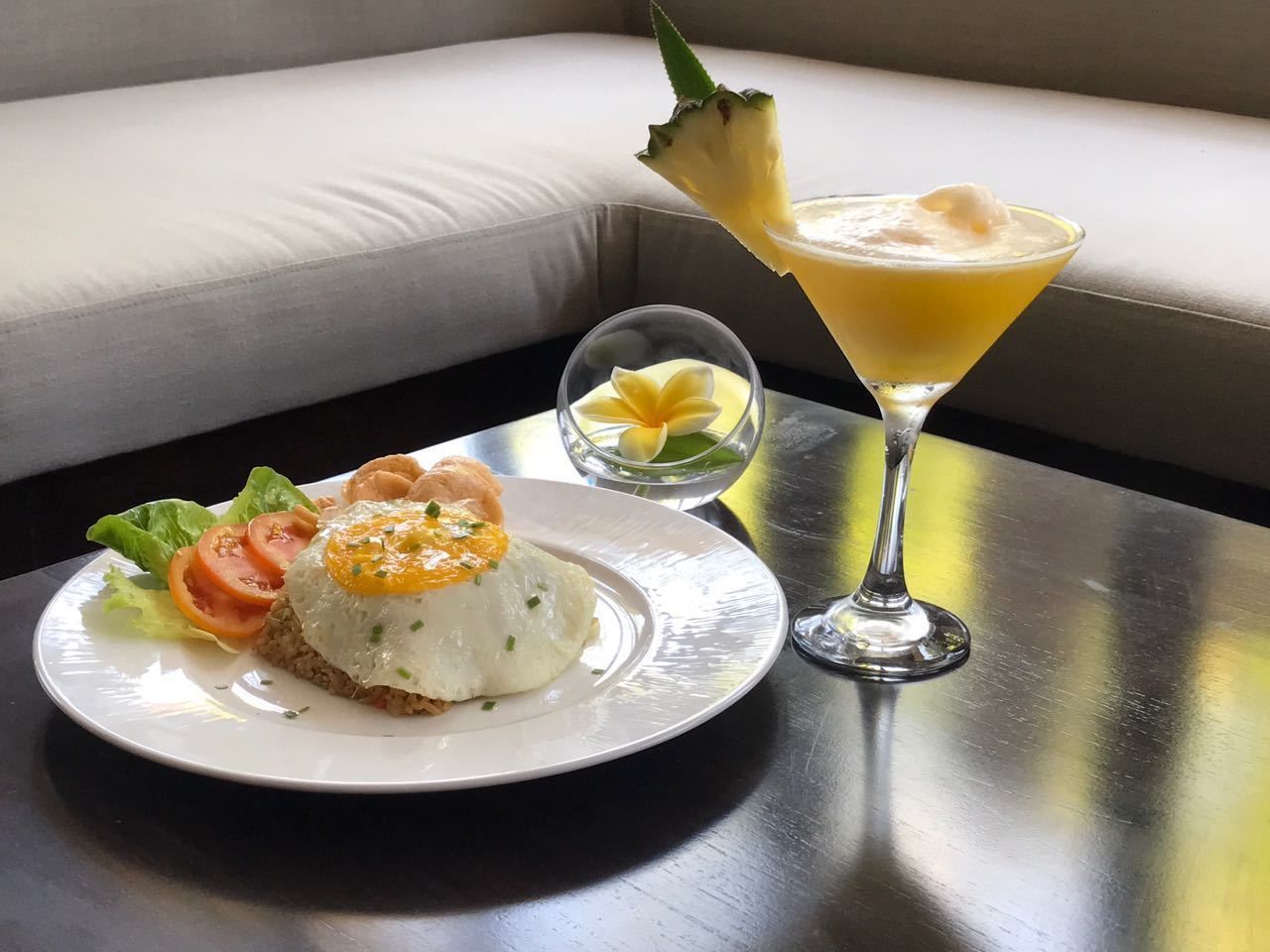 Un plato de comida con un huevo frito encima, un cóctel con una guarnición de piña y un bol pequeño de fruta sobre una mesa.