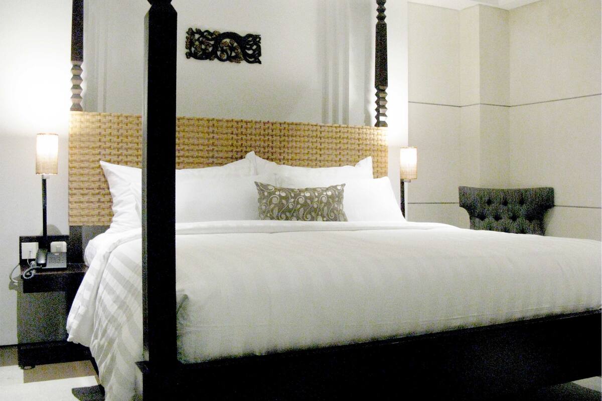 A four-poster bed with a woven headboard, two bedside lamps, and a tufted armchair.