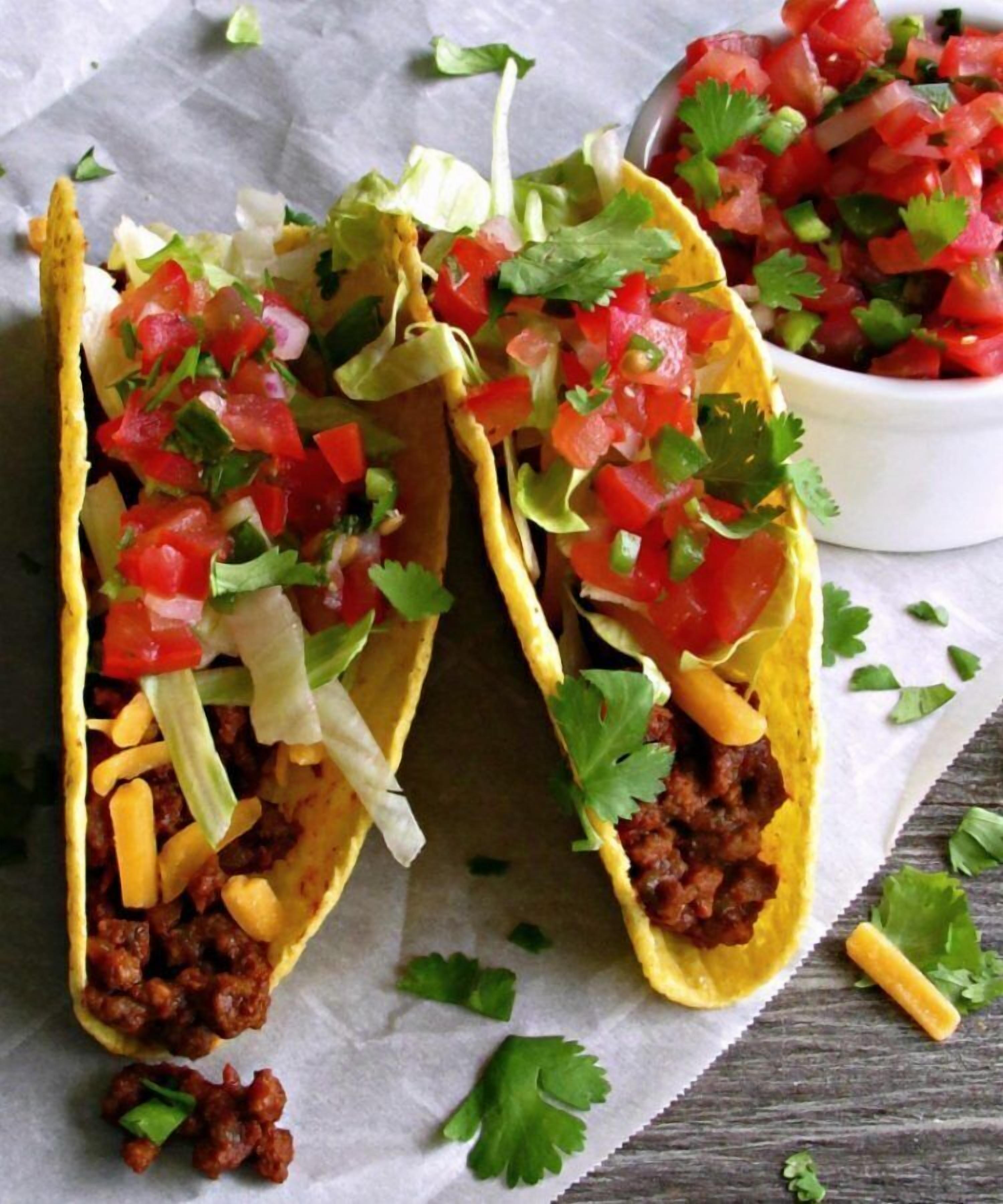 Two tacos with ground meat, cheese, and diced tomatoes on a wooden surface.