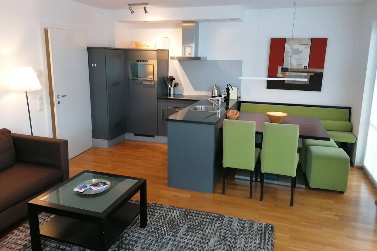 Un espacio habitable compacto con kitchenette, un área de comedor con sillas verdes, un sofá marrón y una mesa de centro de vidrio.