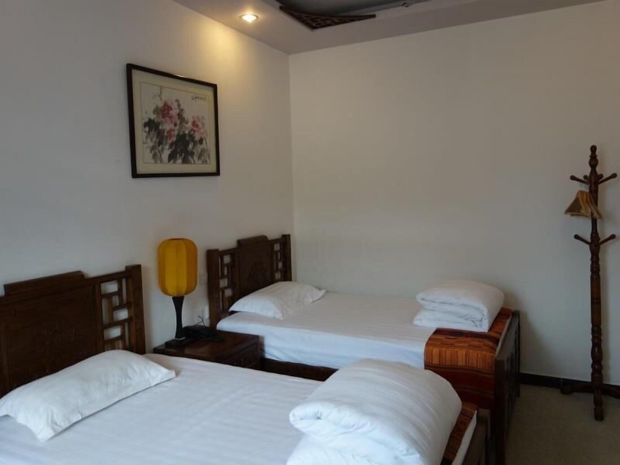 A hotel room with two single beds, a wooden headboard, a bedside lamp, a coat rack, and a framed picture on the wall.