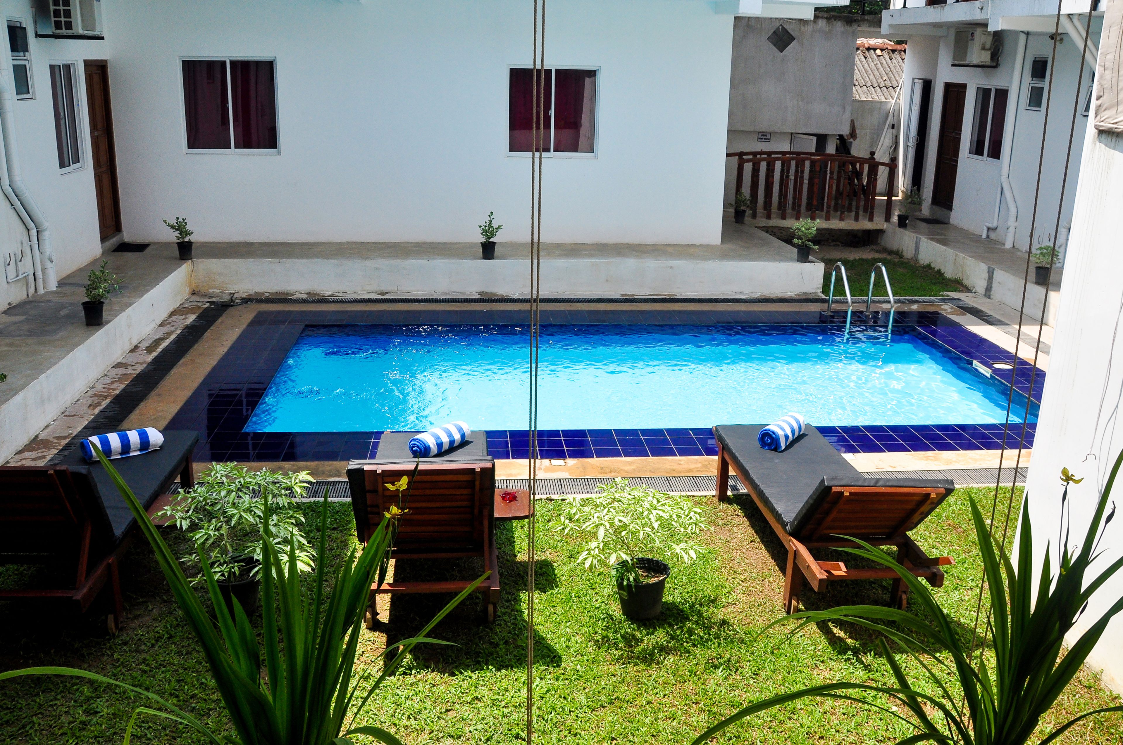 A rectangular swimming pool with a blue interior, surrounded by a white wall. There are two lounge chairs with blue and white striped towels, and several potted plants around the pool area.
