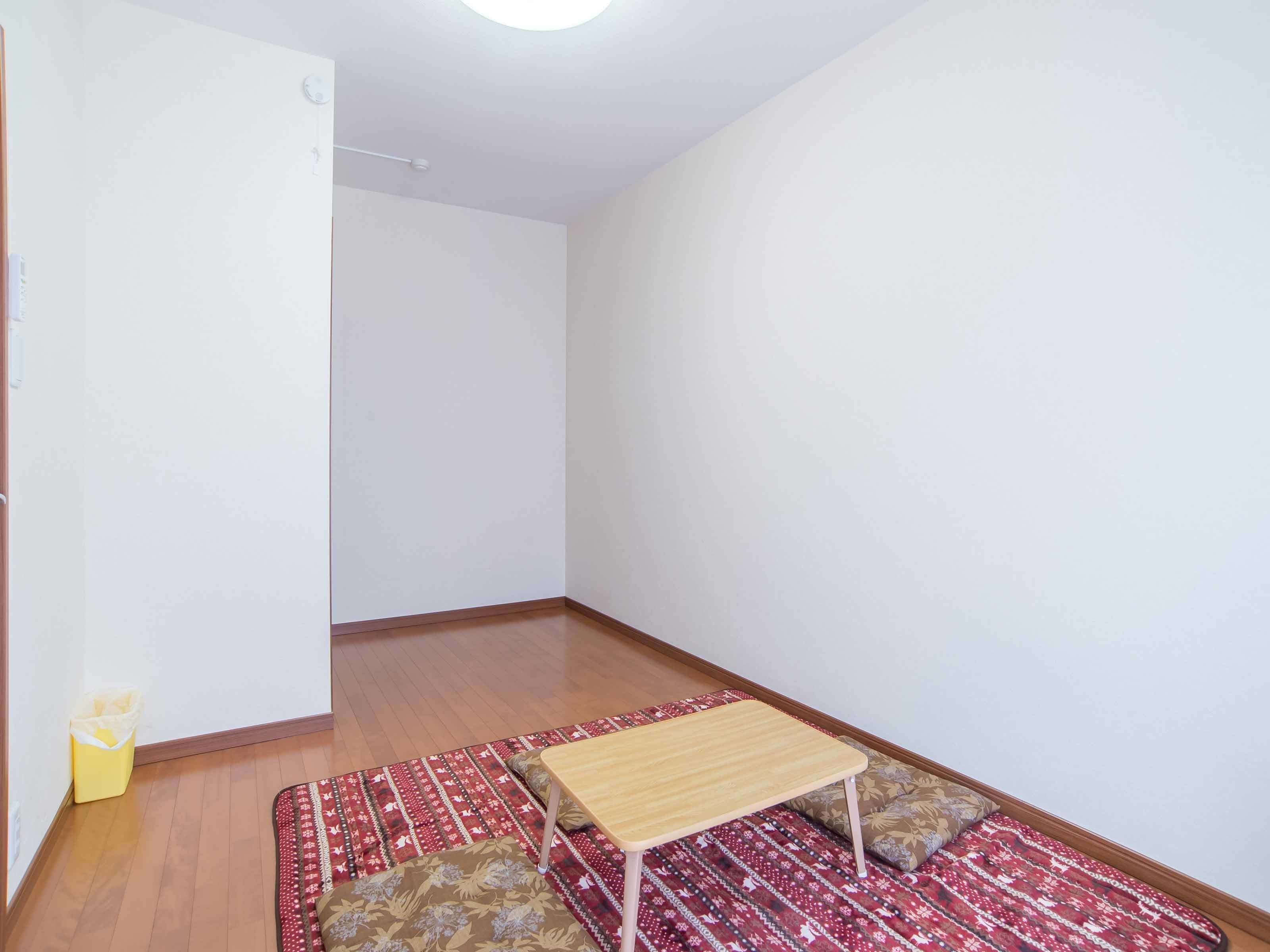 Ein schlichter, spärlich eingerichteter Raum mit Holzboden, einem gemusterten Teppich, einem kleinen Tisch und einem einzelnen Stuhl.