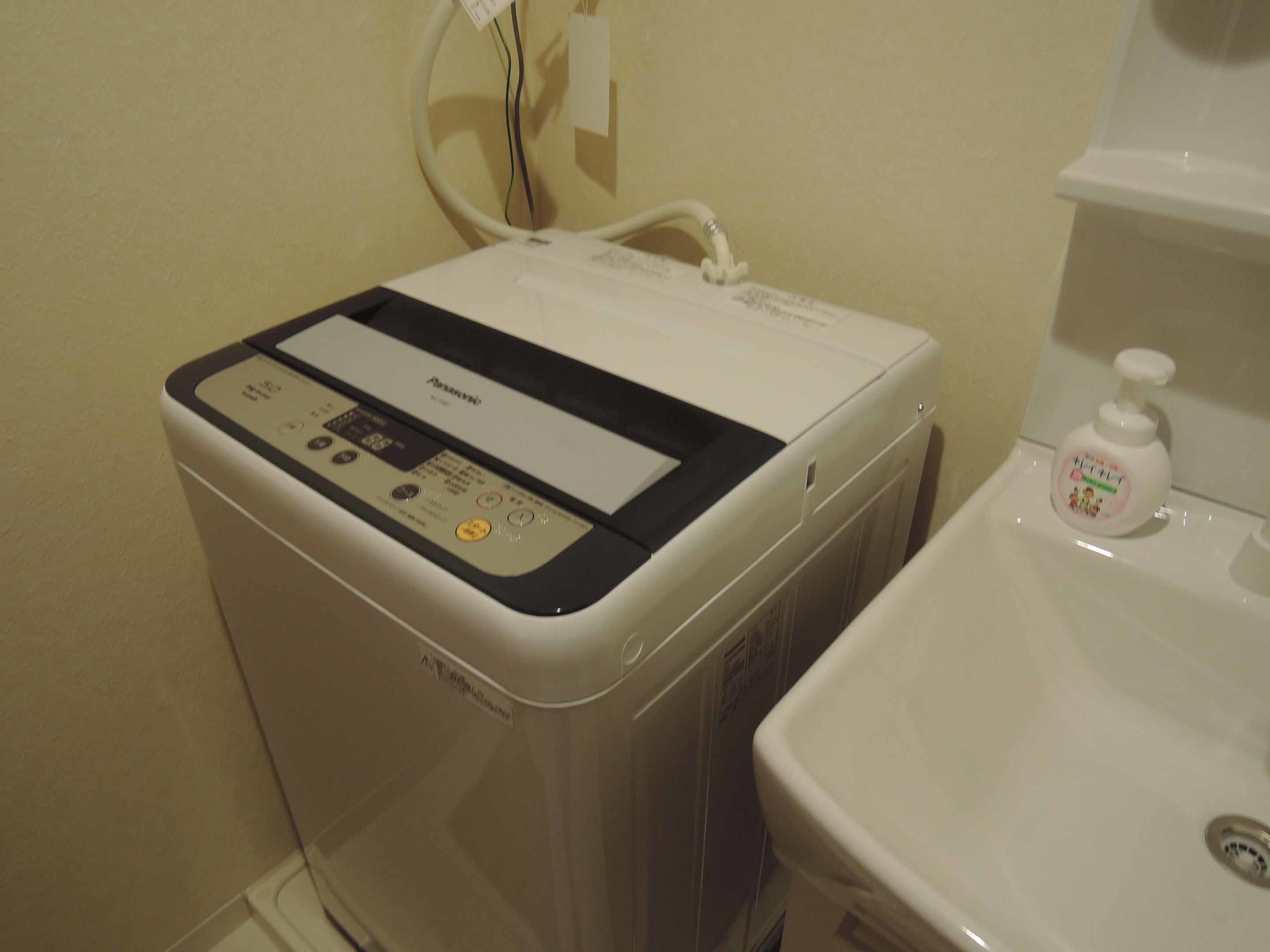 Una lavadora moderna y compacta con panel de control, ubicada junto a un lavamanos con dispensador de jabón.