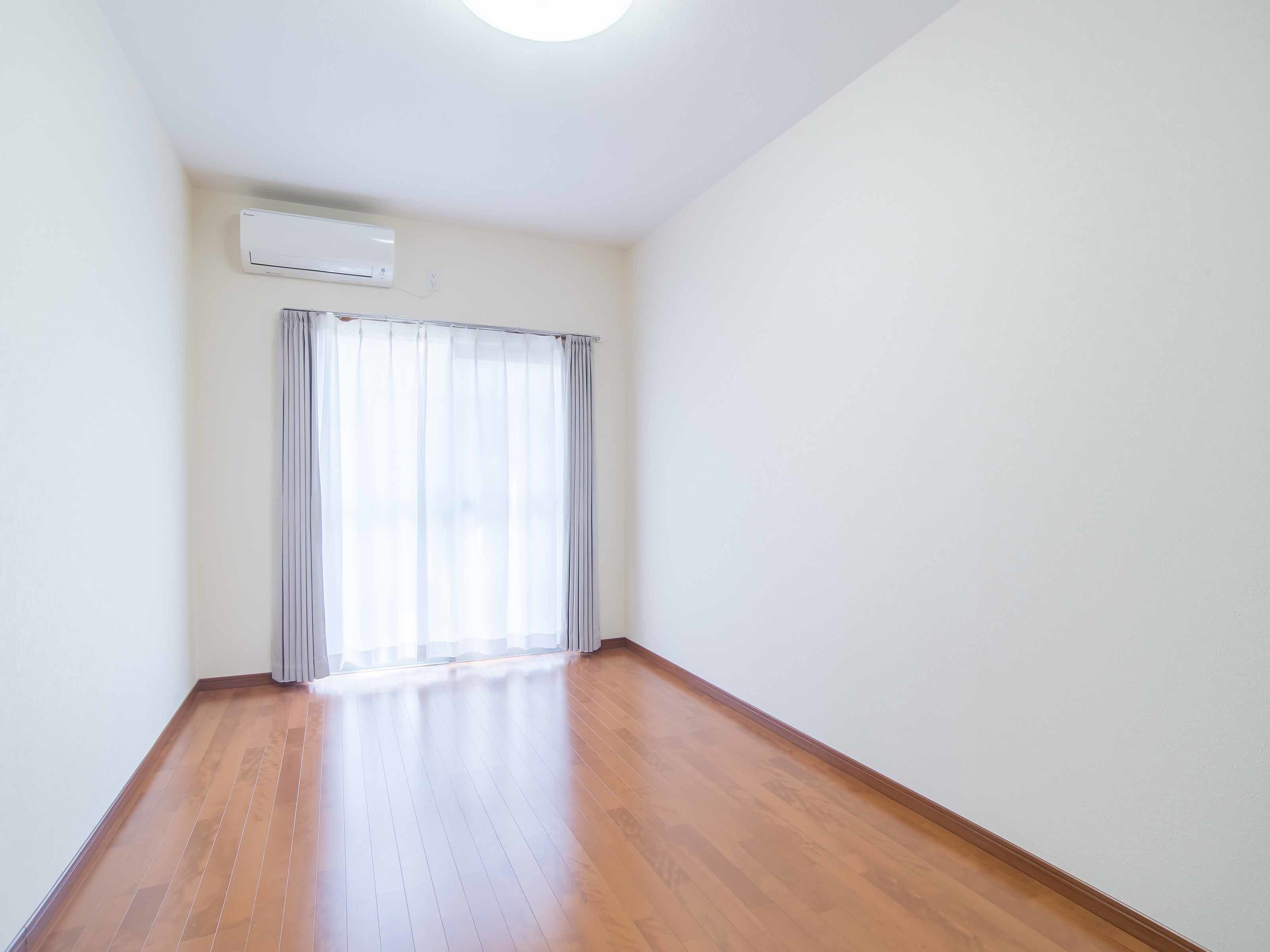 Una habitación con piso de madera, una ventana con cortinas translúcidas y un equipo de aire acondicionado en la pared.