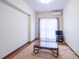 Una habitación estrecha con una mesa de vidrio, un televisor sobre un mueble bajo y una ventana con cortinas.