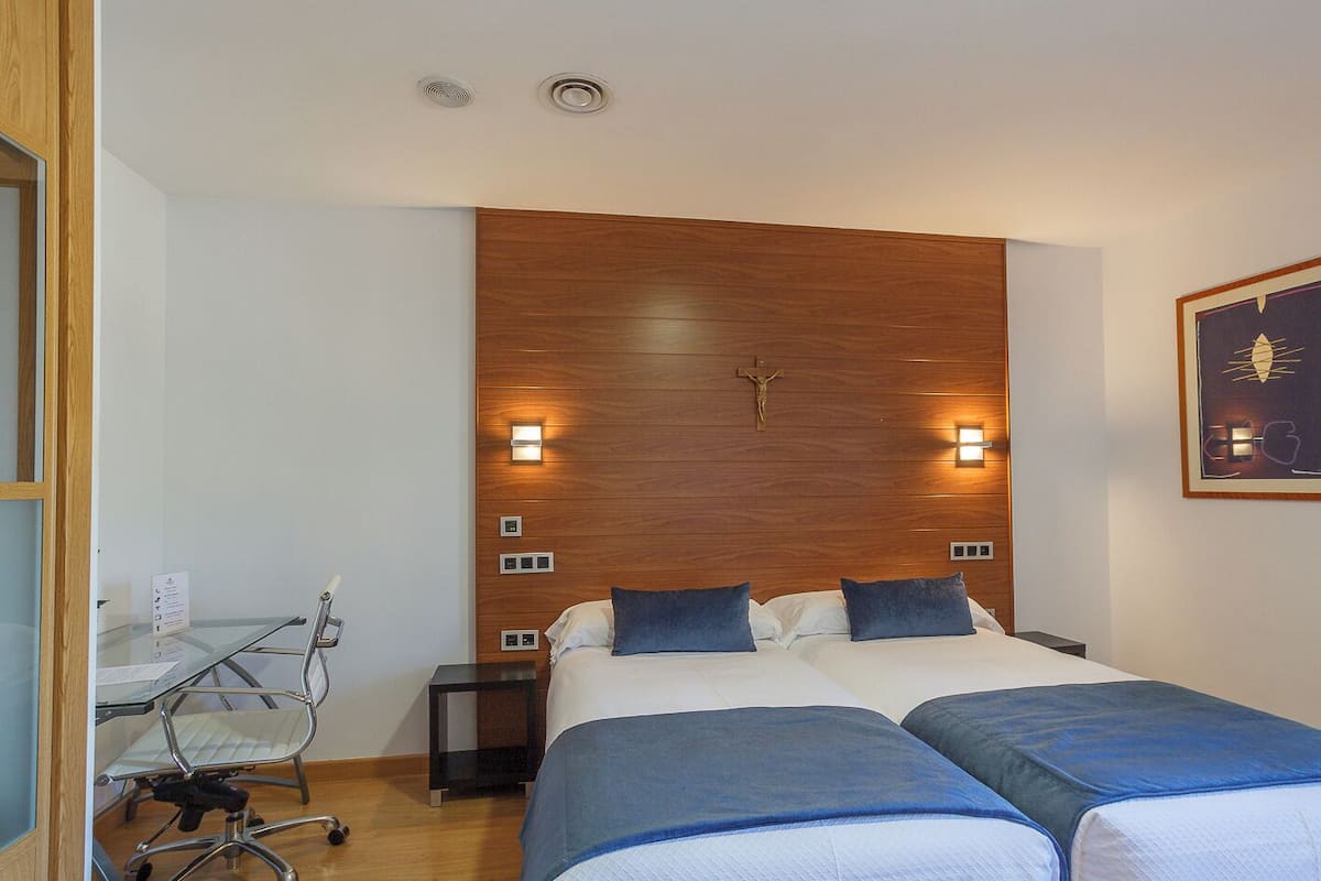 A hotel room with a wooden headboard, two beds with blue bedding, a desk with a chair, and a wall-mounted light fixture.