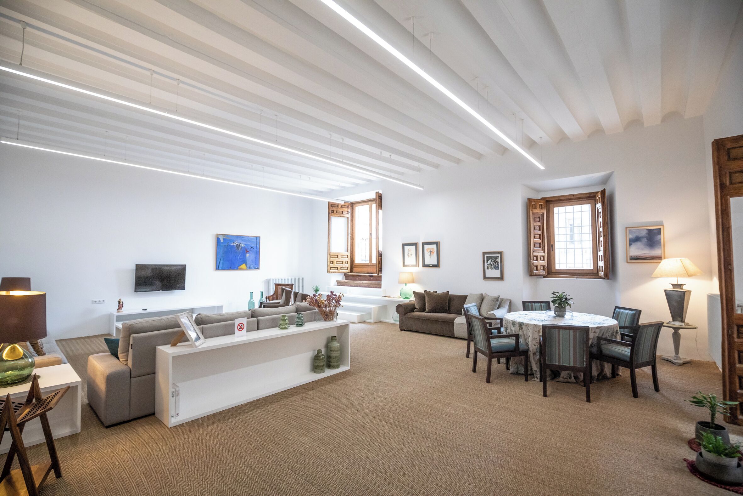 Una sala de estar moderna con un sofá, una mesa de comedor y sillas. Hay un televisor en la pared y una ventana con contraventanas de madera.