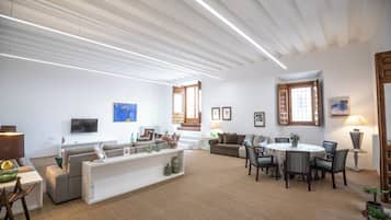 Una sala de estar moderna con un sofá, una mesa de comedor y sillas. Hay un televisor en la pared y una ventana con contraventanas de madera.