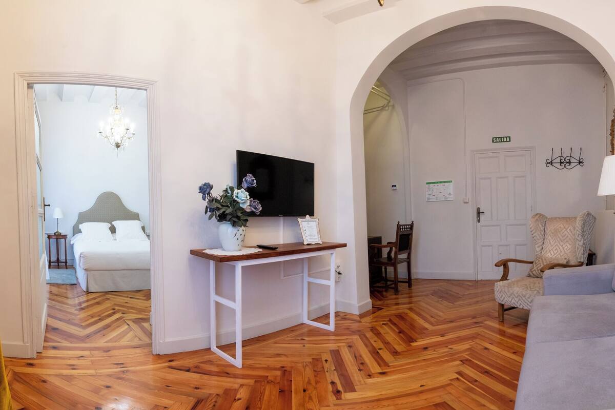 A hotel room with a wooden floor, a bed, a desk with a TV, a sofa, and an arched doorway leading to another room.