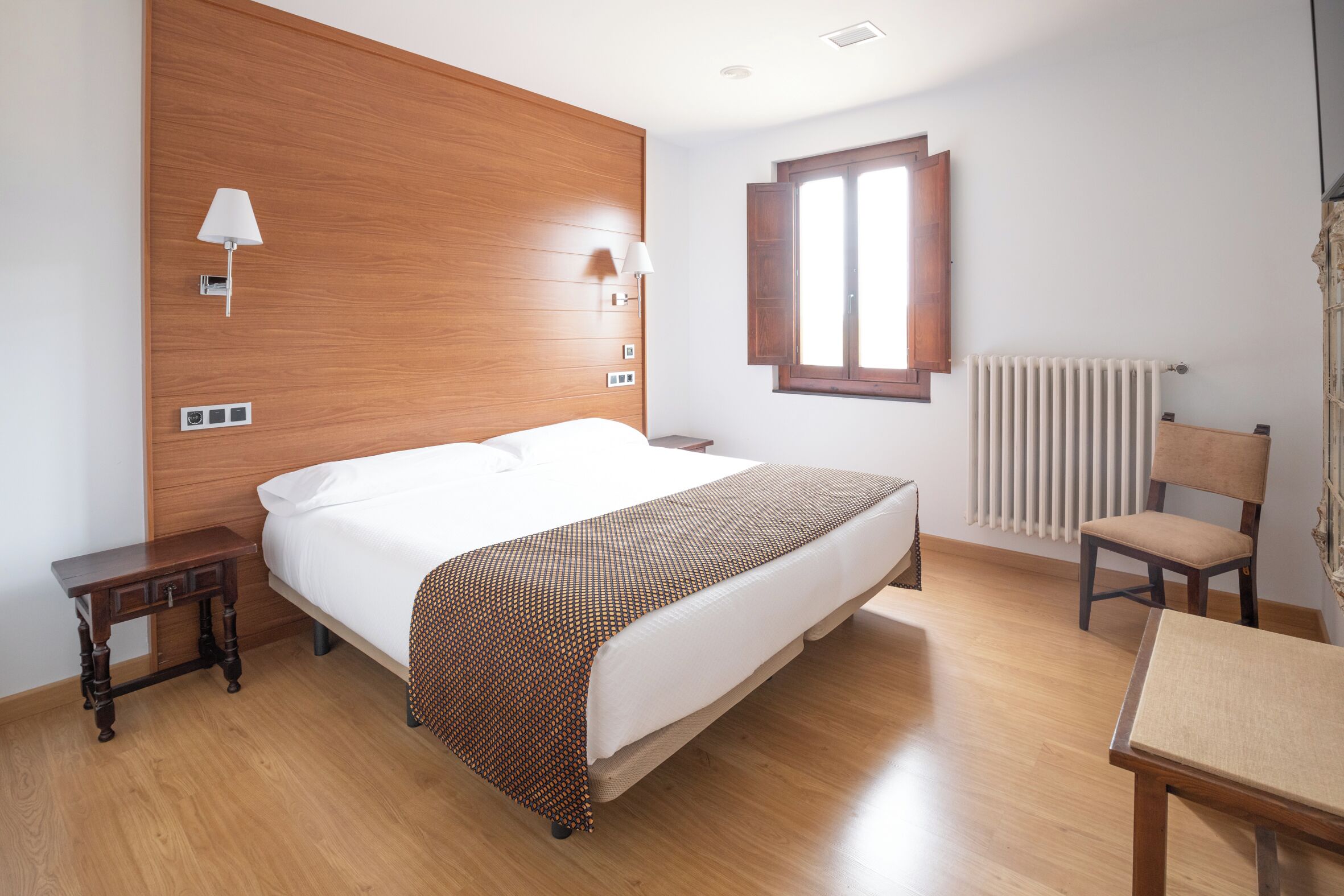 Habitación de hotel con una cama grande, cabecera de madera, mesitas de noche, una silla, un radiador y una ventana.