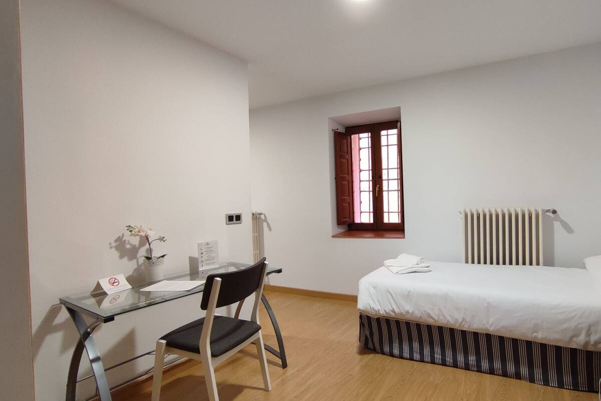 A bedroom with a bed, a desk with a chair, a radiator, and a window with wooden shutters.