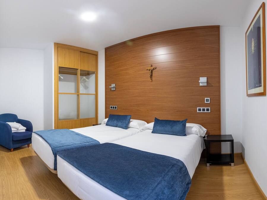 Un dormitorio con dos camas, cabecero de madera, una silla azul, una mesita de noche y un cuadro enmarcado en la pared.