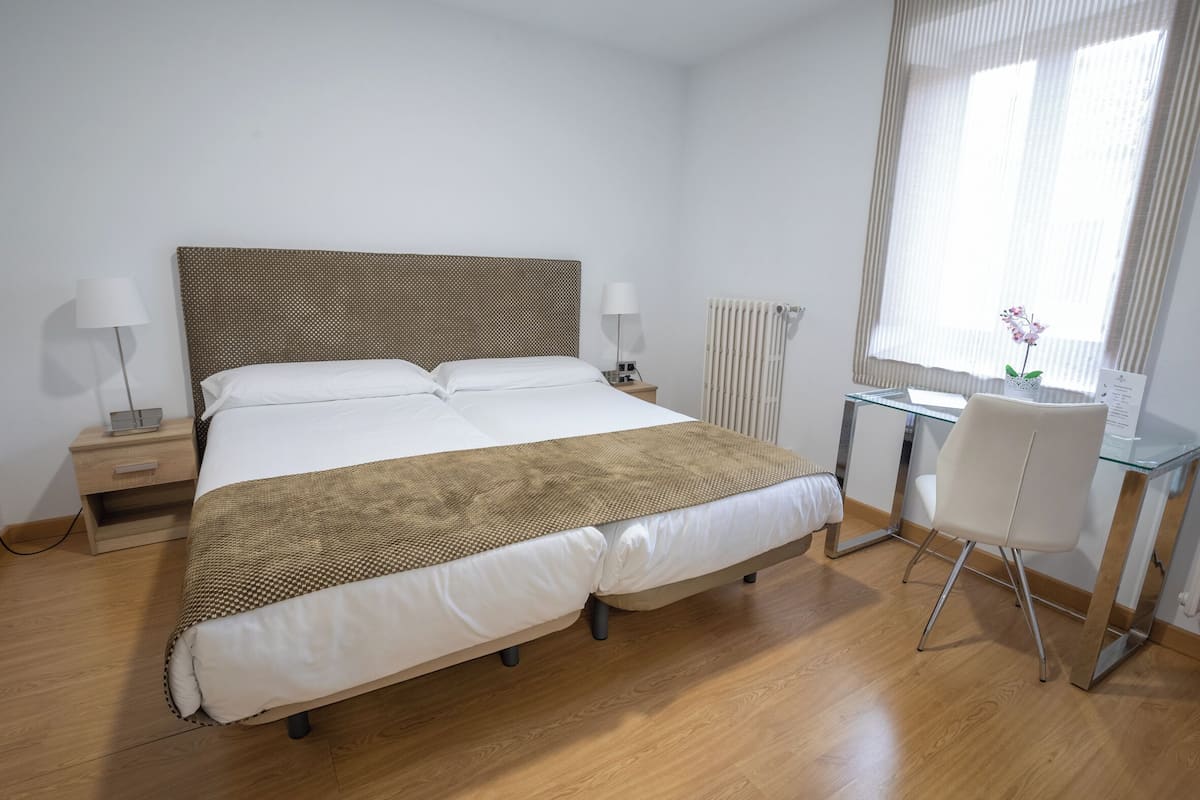 A neatly arranged bedroom with a large bed, bedside tables with lamps, a desk with a chair, and a window with curtains.