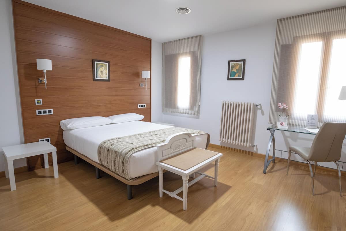 A hotel room with a wooden headboard, a bed with white linens, a small table, a chair, a desk with a computer, and a window with curtains.