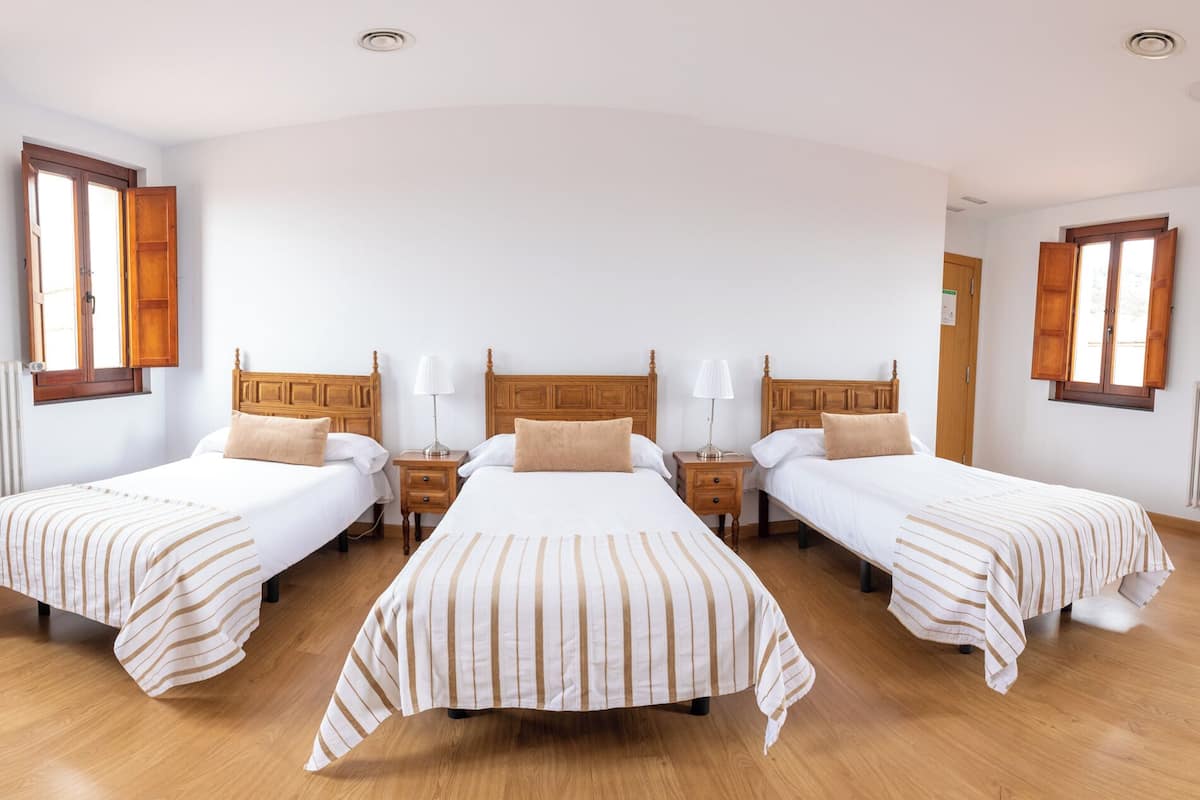 A hotel room with three single beds, wooden headboards, white bedding, and a wooden floor.