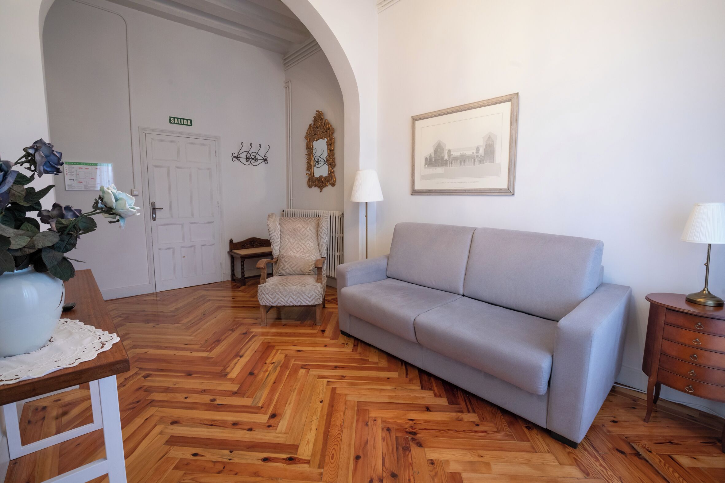 A room with a herringbone wooden floor, a grey sofa, a patterned armchair, a wooden side table, a white vase with flowers, a framed picture on the wall, and a door with a sign.