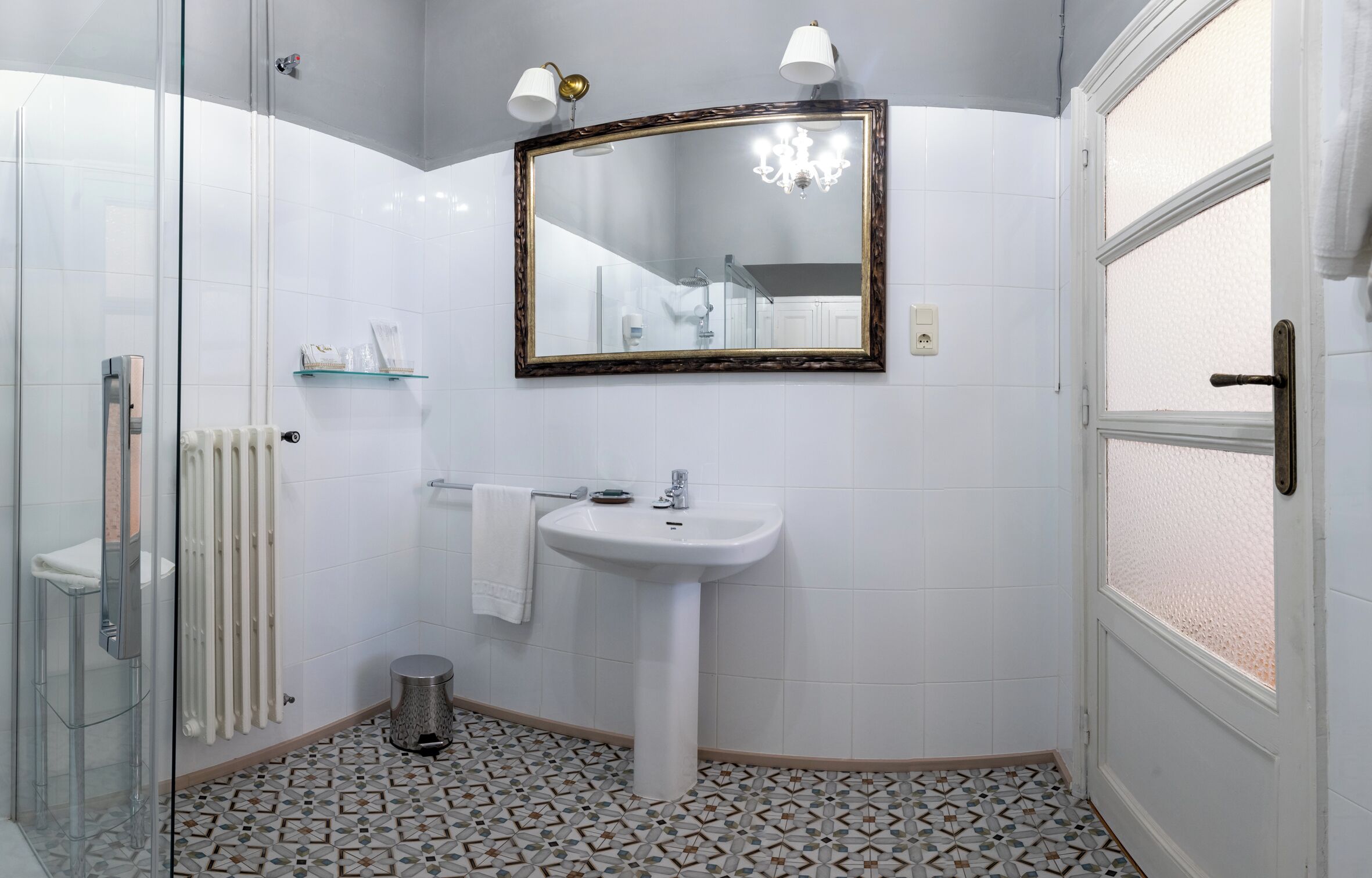 Un bany amb un lavabo blanc, una cabina de dutxa de vidre, un sòl amb dibuixos i un mirall amb marc decoratiu.