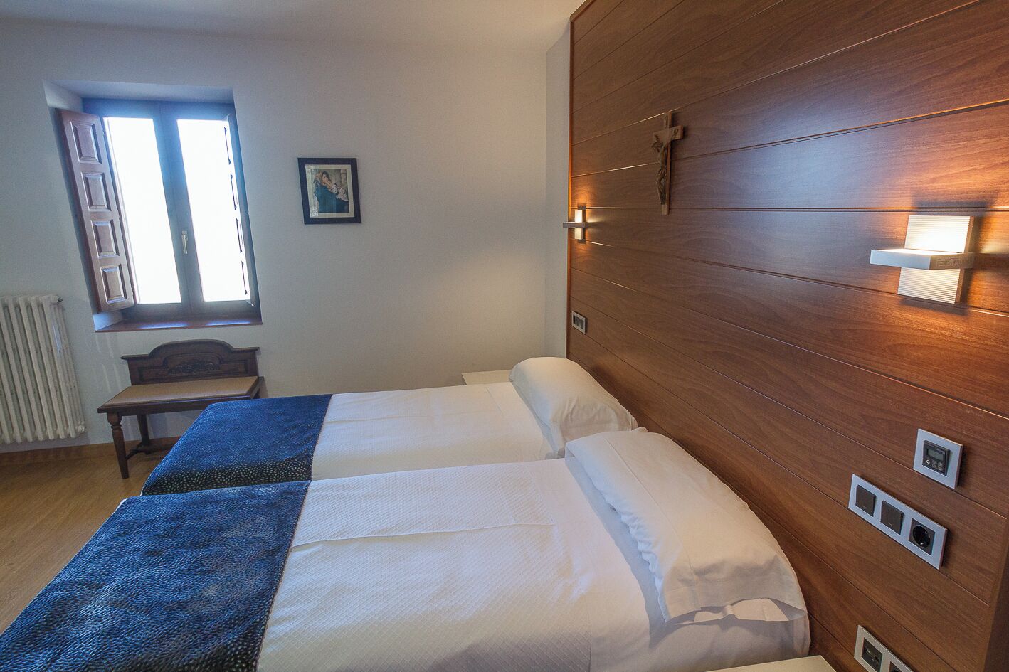 Habitación de hotel con dos camas, cabecero de madera, ventana con persianas, radiador y un cuadro en la pared.