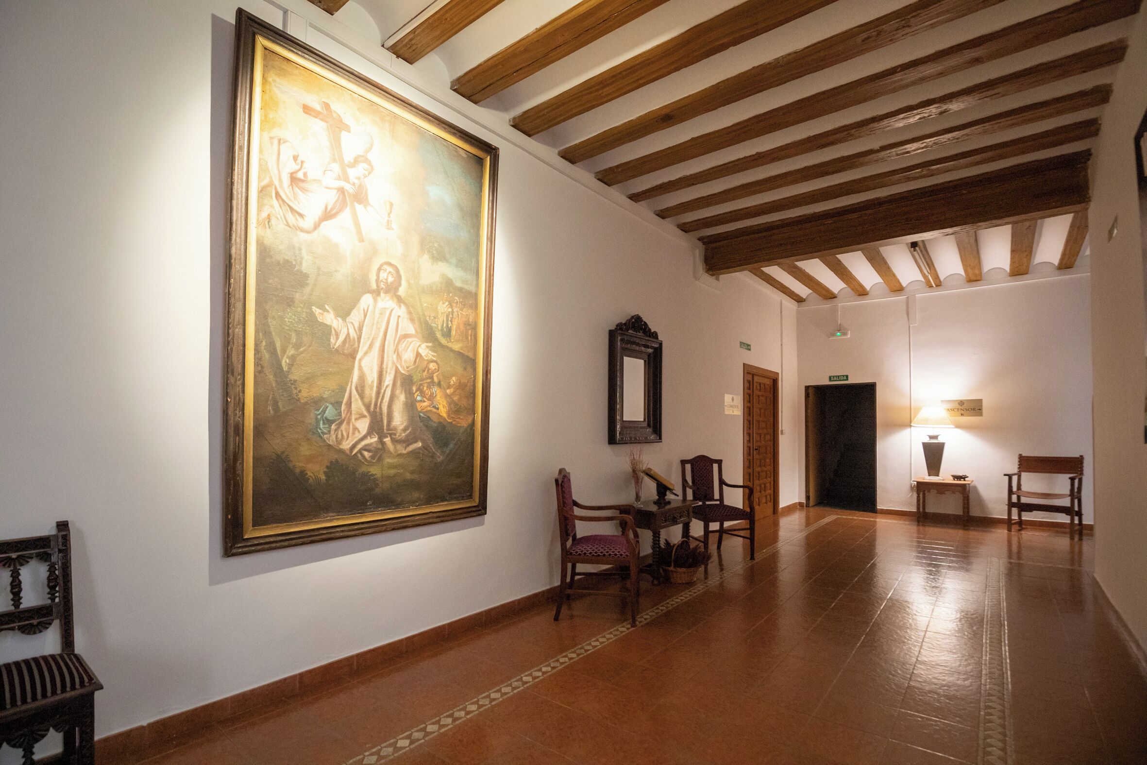 Un pasillo con un cuadro religioso grande, vigas de madera y un banco.