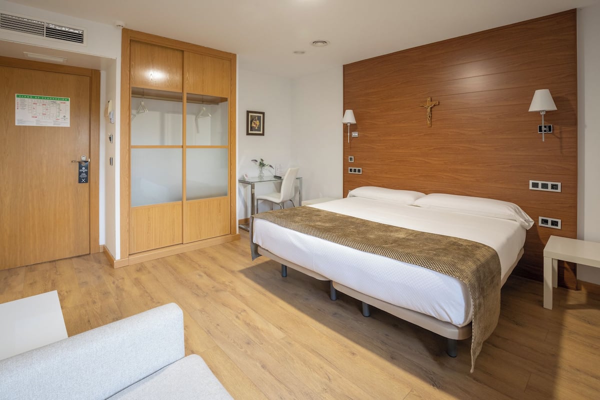 A hotel room with a wooden wardrobe, a bed with a white mattress and a brown blanket, a small table with a lamp, and a wall-mounted light fixture.