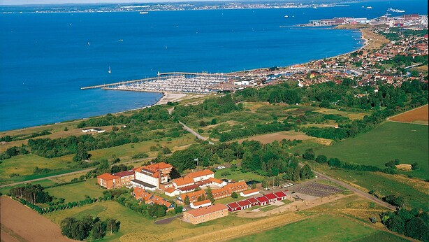 Luchtbeeld van een kustplaatsje met een jachthaven, groene velden en een groot complex met rode daken.