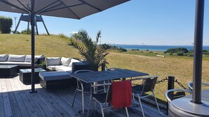 Een terras met buitenmeubilair, waaronder een bank, stoelen en een tafel, met uitzicht op een gazon en de zee.