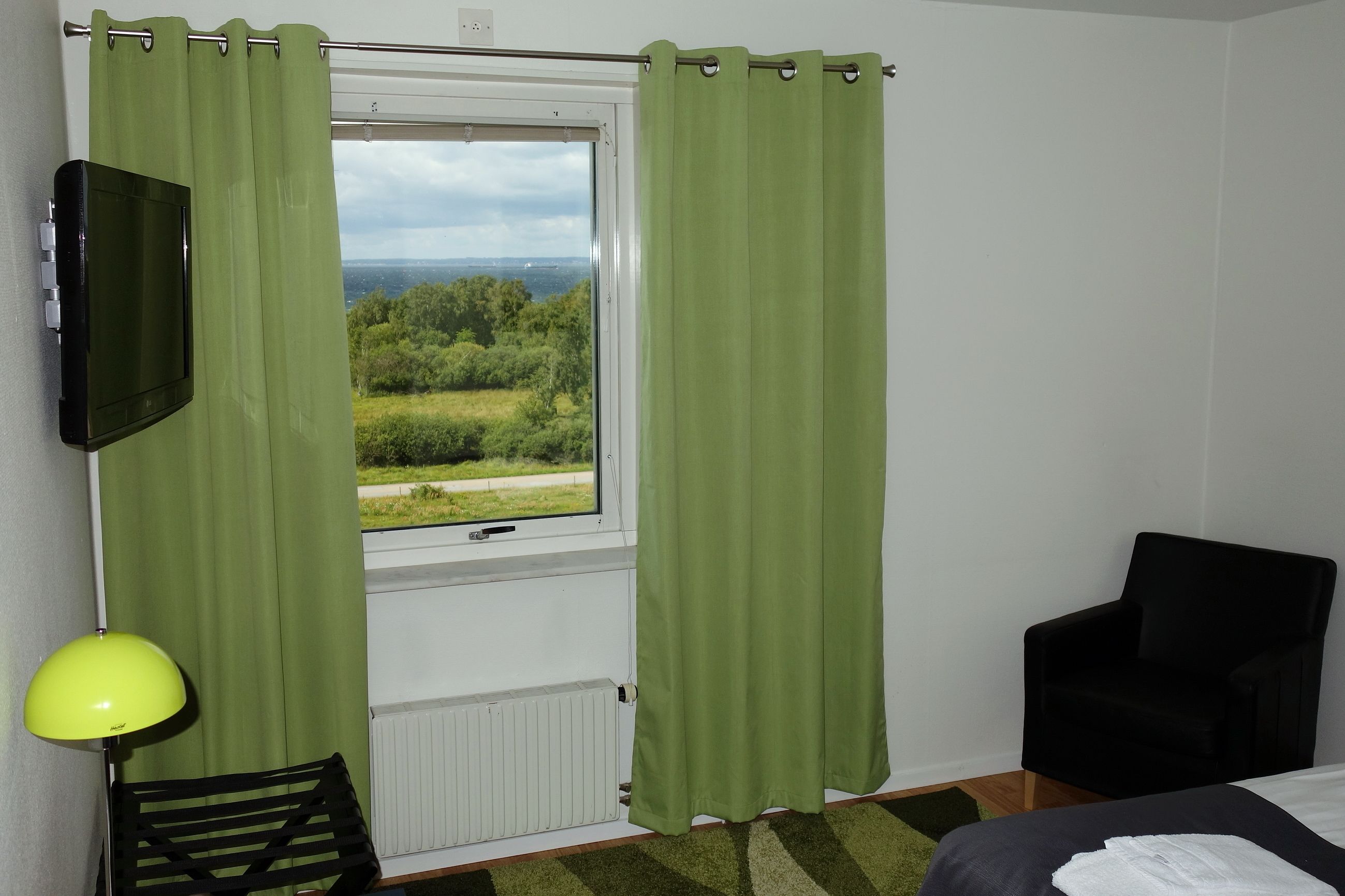 Una habitación con ventana, televisor montado en la pared, una silla y una cama.