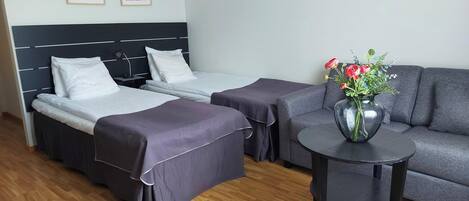 Habitación de hotel con dos camas, un sofá gris y una mesita con un jarrón de flores.