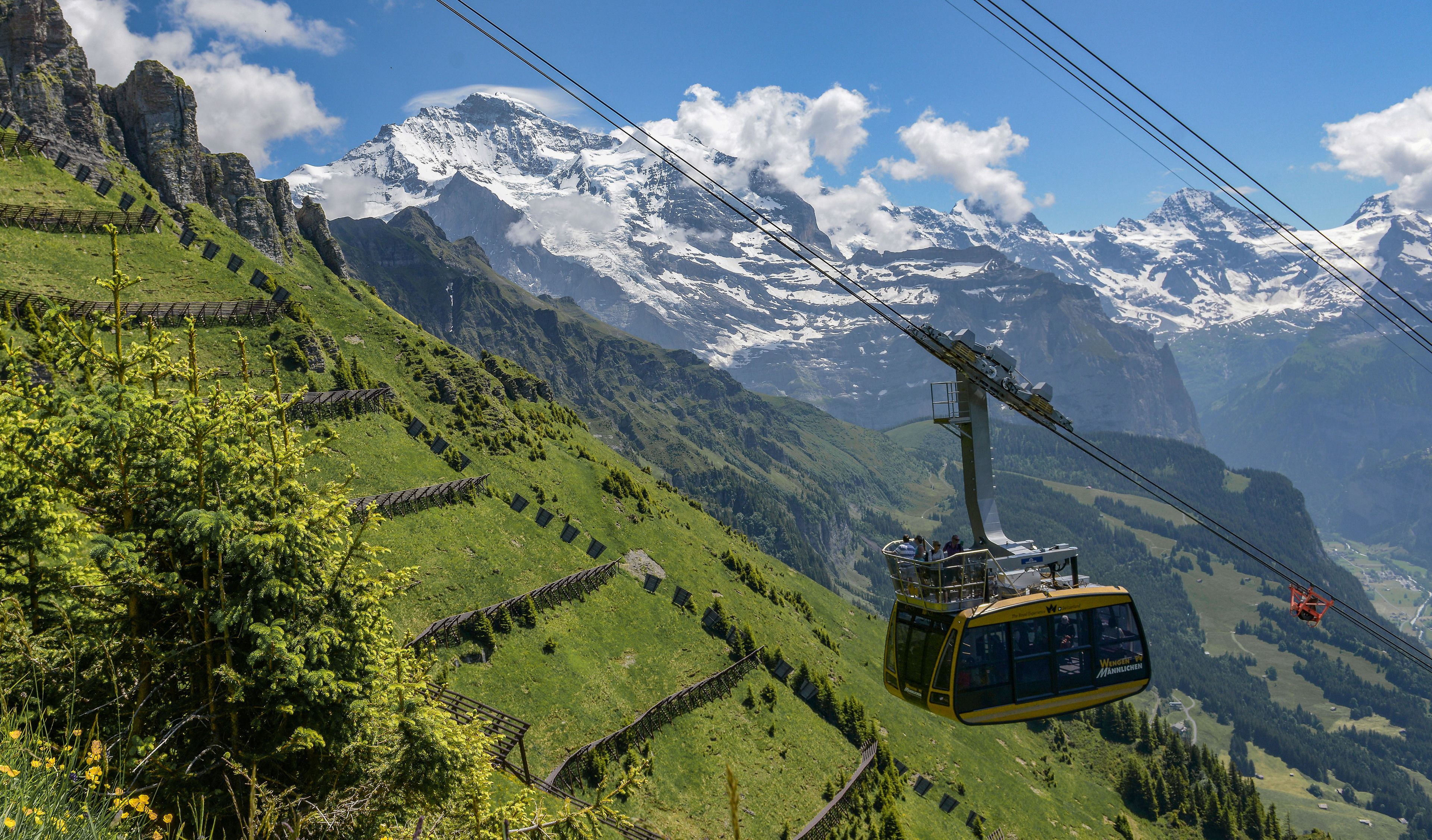 Eine Seilbahn fährt einen Berg hinauf, im Hintergrund schneebedeckte Gipfel.