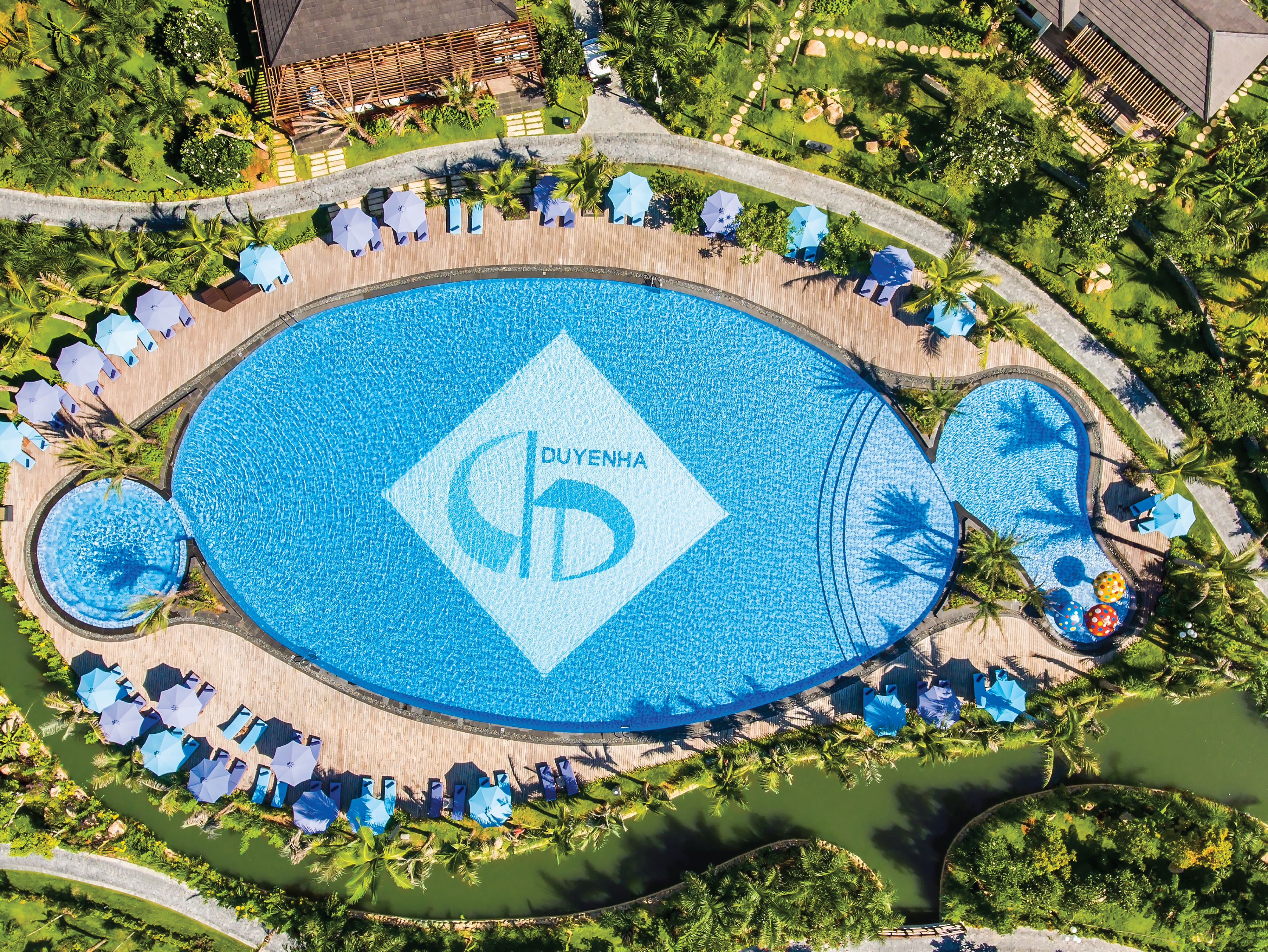 Een overzicht vanuit de lucht van een resort met een groot centraal zwembad, een houten terras eromheen en palmbomen.