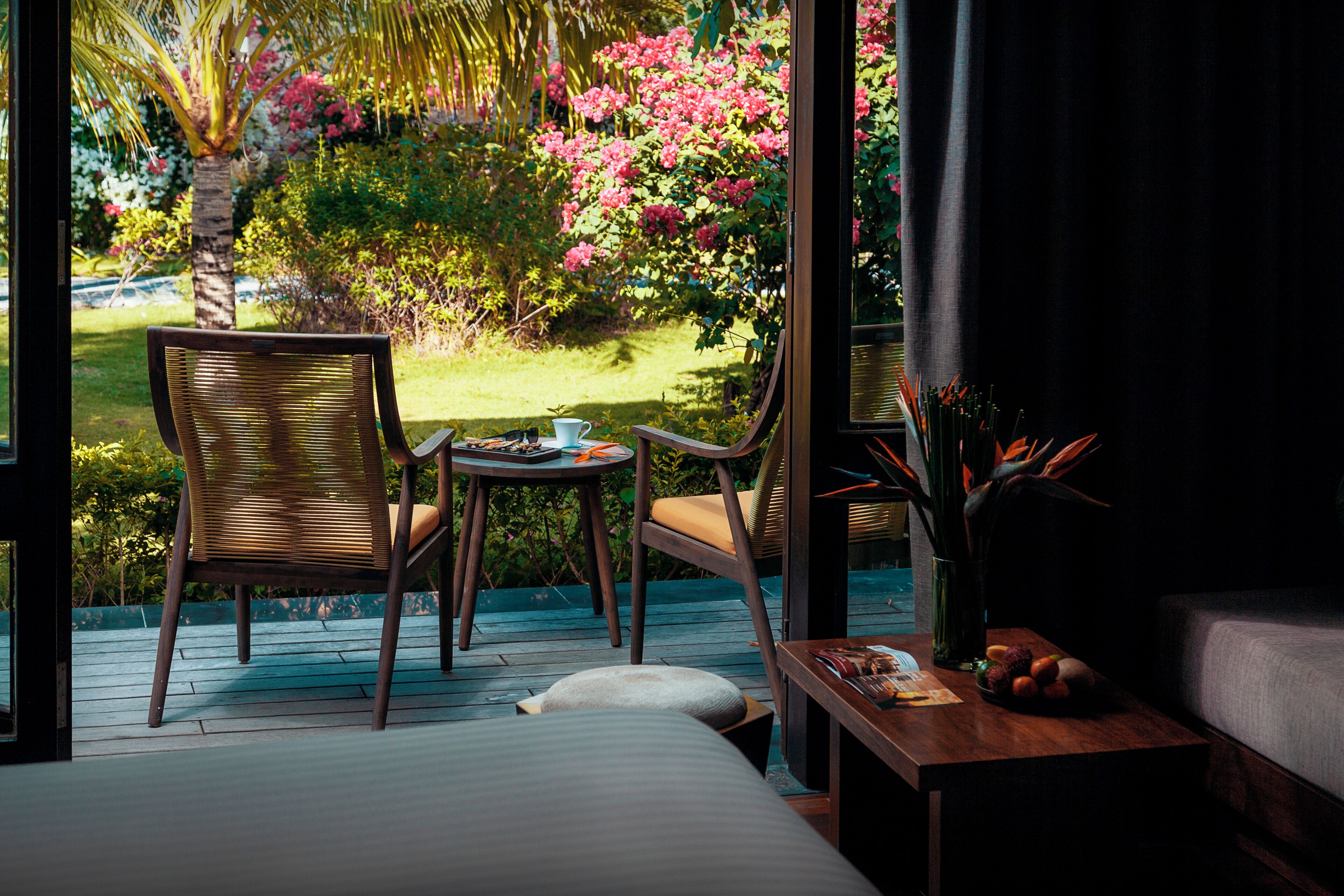 Una habitación con una cama, una mesa de madera con un cuenco de fruta, una silla de mimbre y vistas a un jardín con flores rosas y una palmera.
