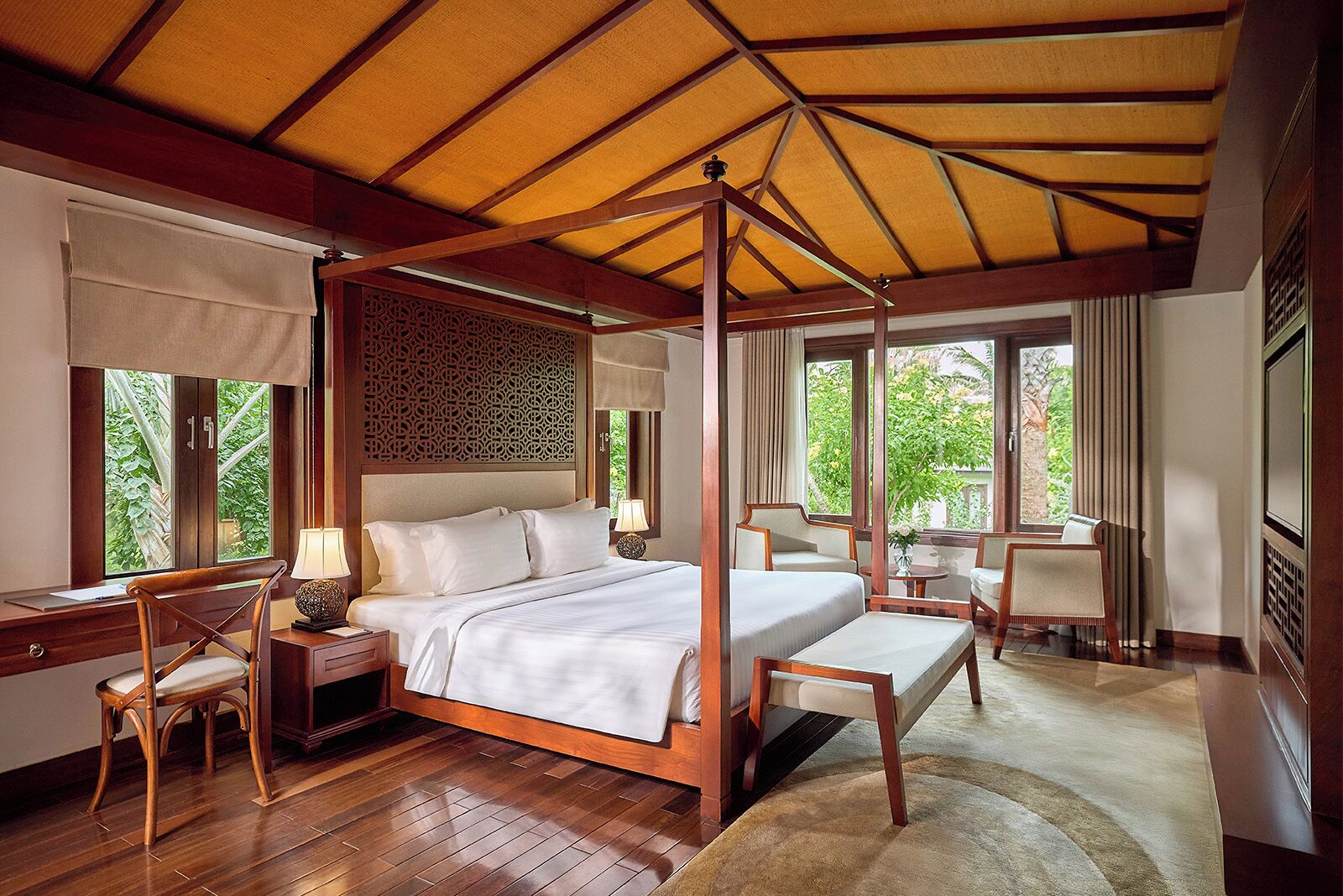 A four-poster bed with a canopy, a desk with a chair, and a view of greenery outside.