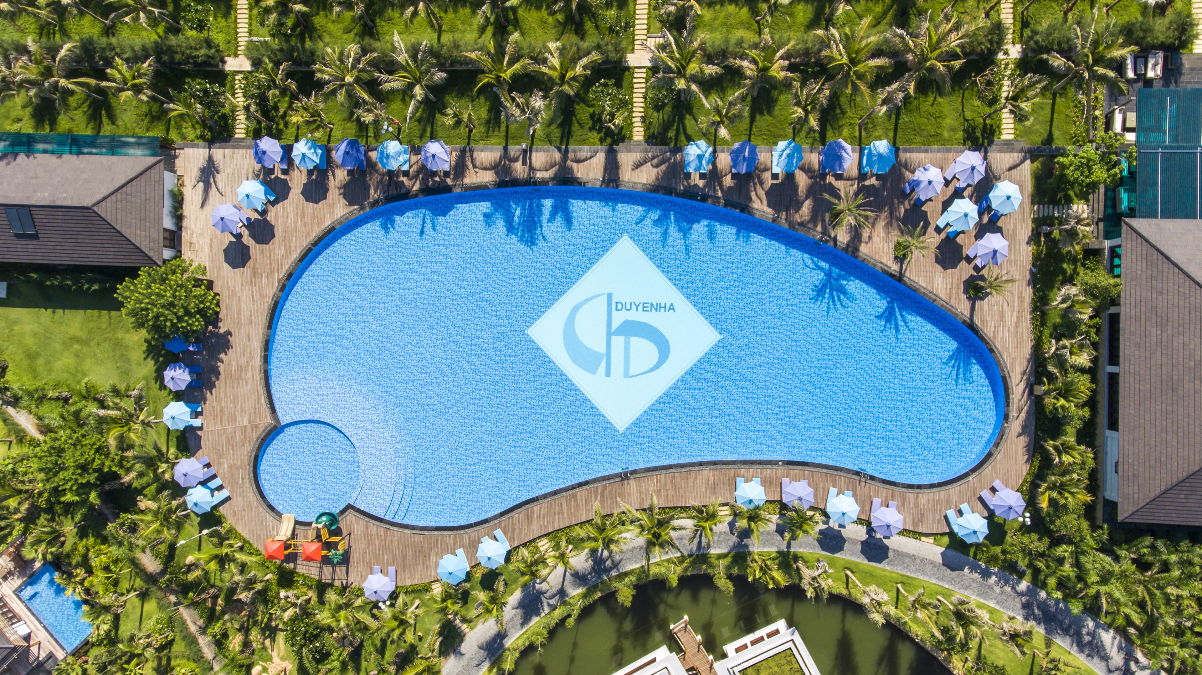 An aerial view of a resort with a large swimming pool, palm trees, and a wooden deck with umbrellas.