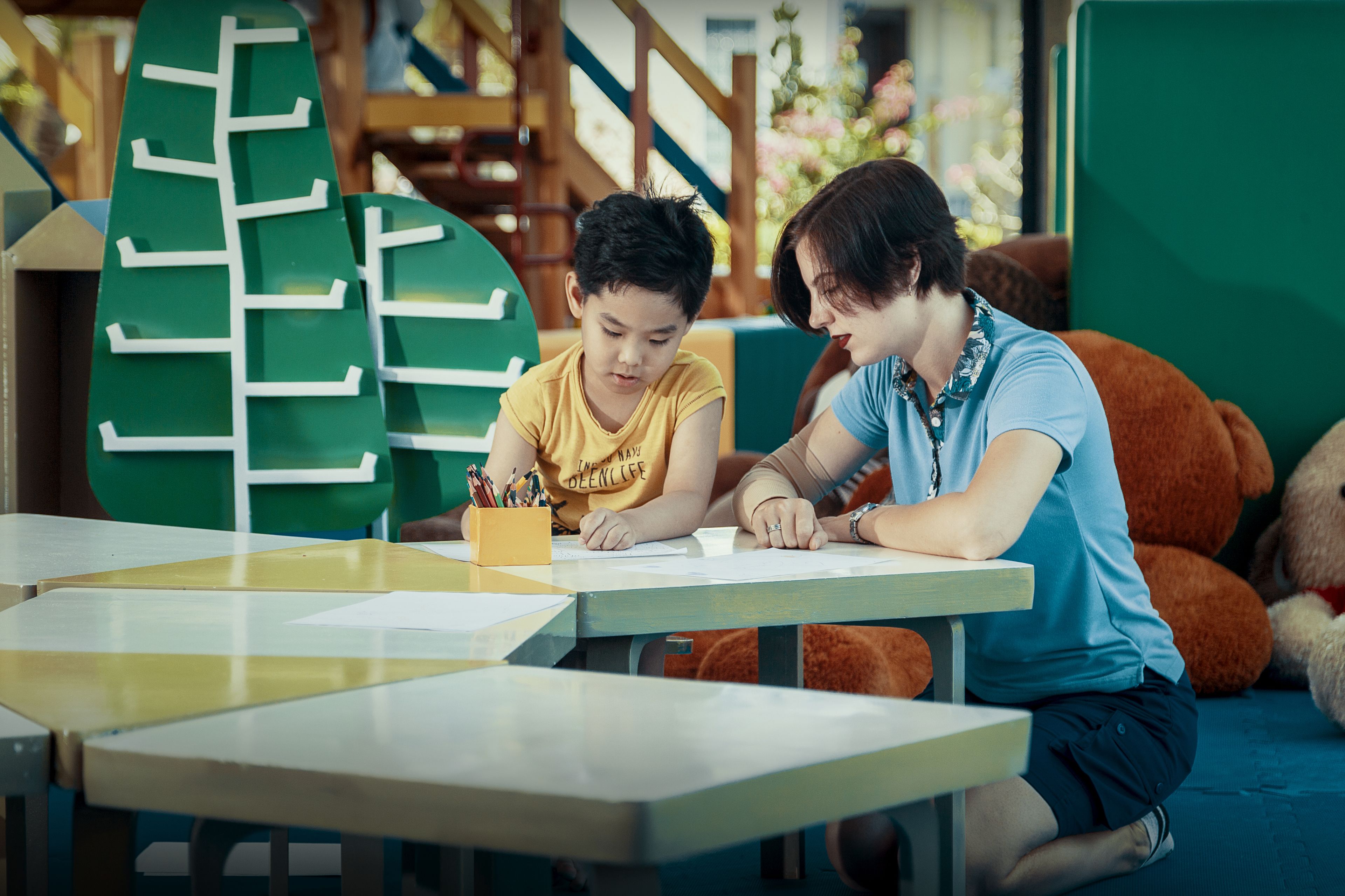 Un enfant et un adulte sont assis à une table, devant une décoration verte en forme d’arbre, un ours en peluche et un porte-crayons jaune.