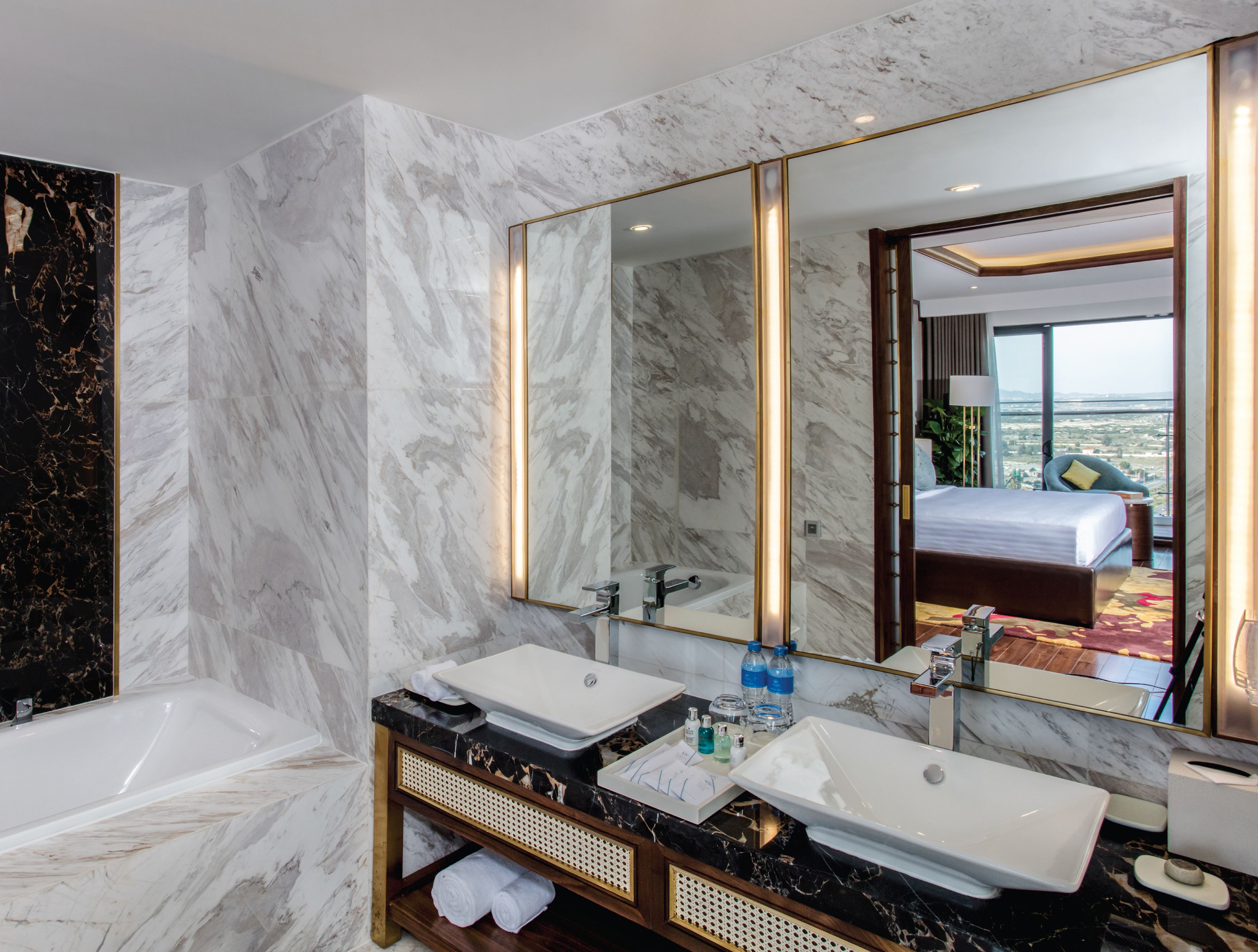A modern bathroom with marble walls, a large mirror, two sinks, a bathtub, and a view of a bedroom through the mirror.