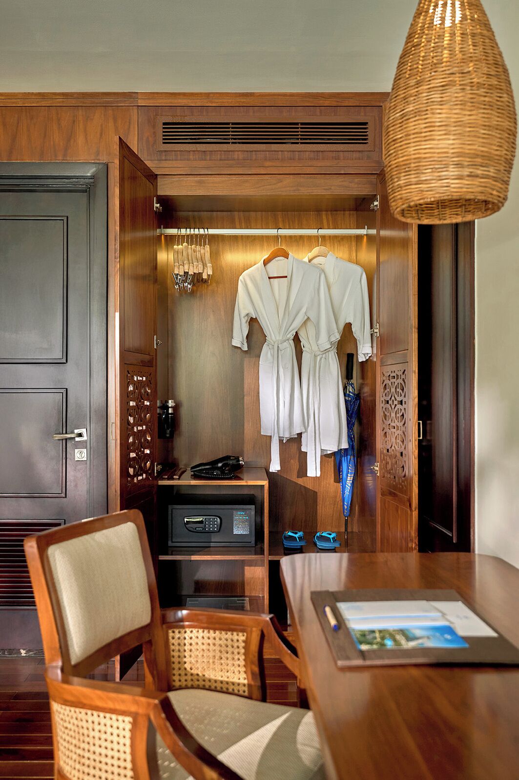 A hotel room with a wooden wardrobe, a robe and towel rack, a desk with a telephone and a digital device, and a wicker lamp.