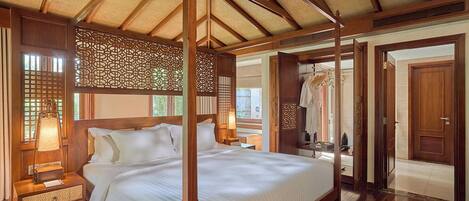A traditional wooden four-poster bed with a woven ceiling, a nightstand with a lamp, and a view of a clothing rack and door.
