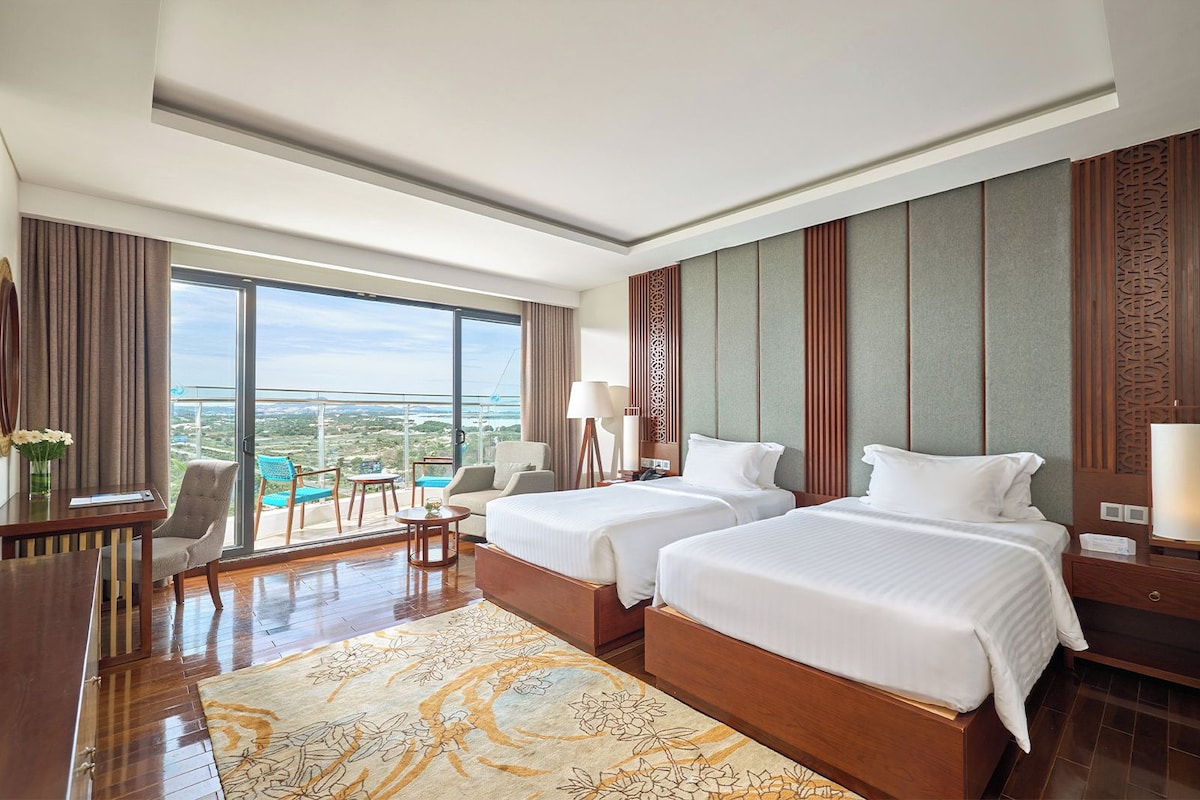 A hotel room with two beds, a balcony with a view, a desk, and a chair.