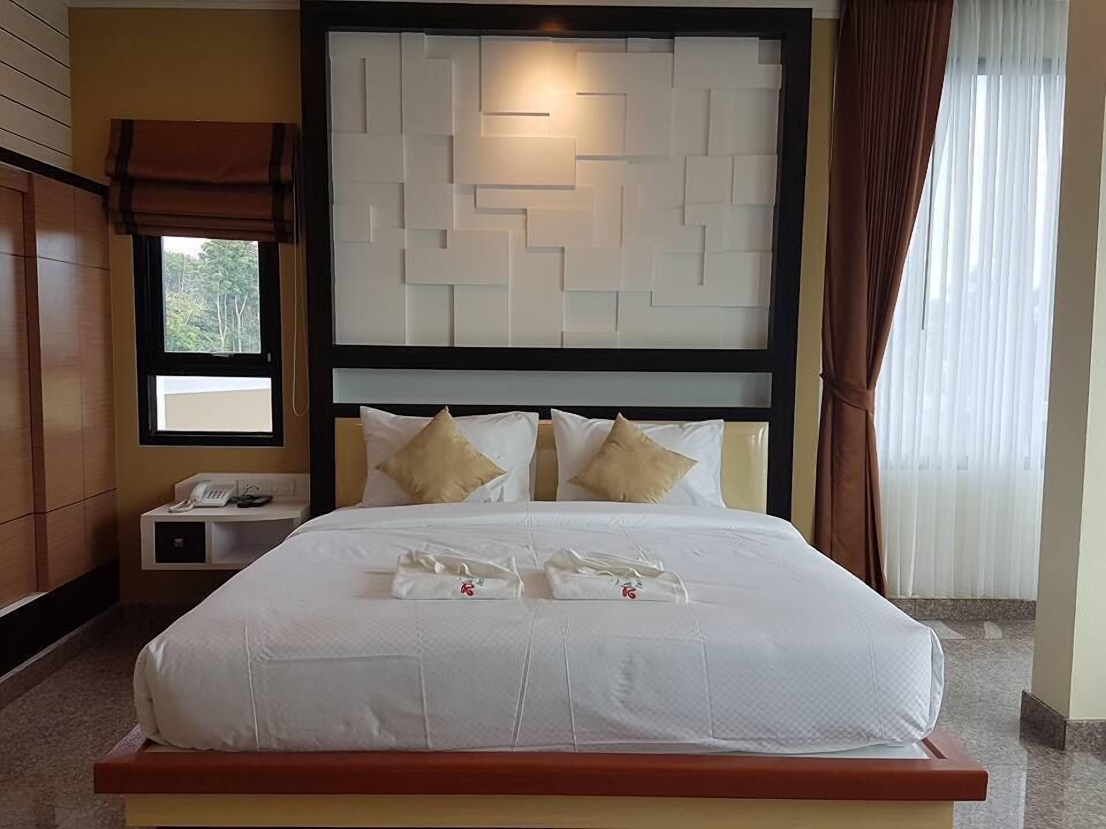 A neatly made bed with white linens and two decorative pillows, a bedside table with a phone and a lamp, and a window with a view of greenery outside.