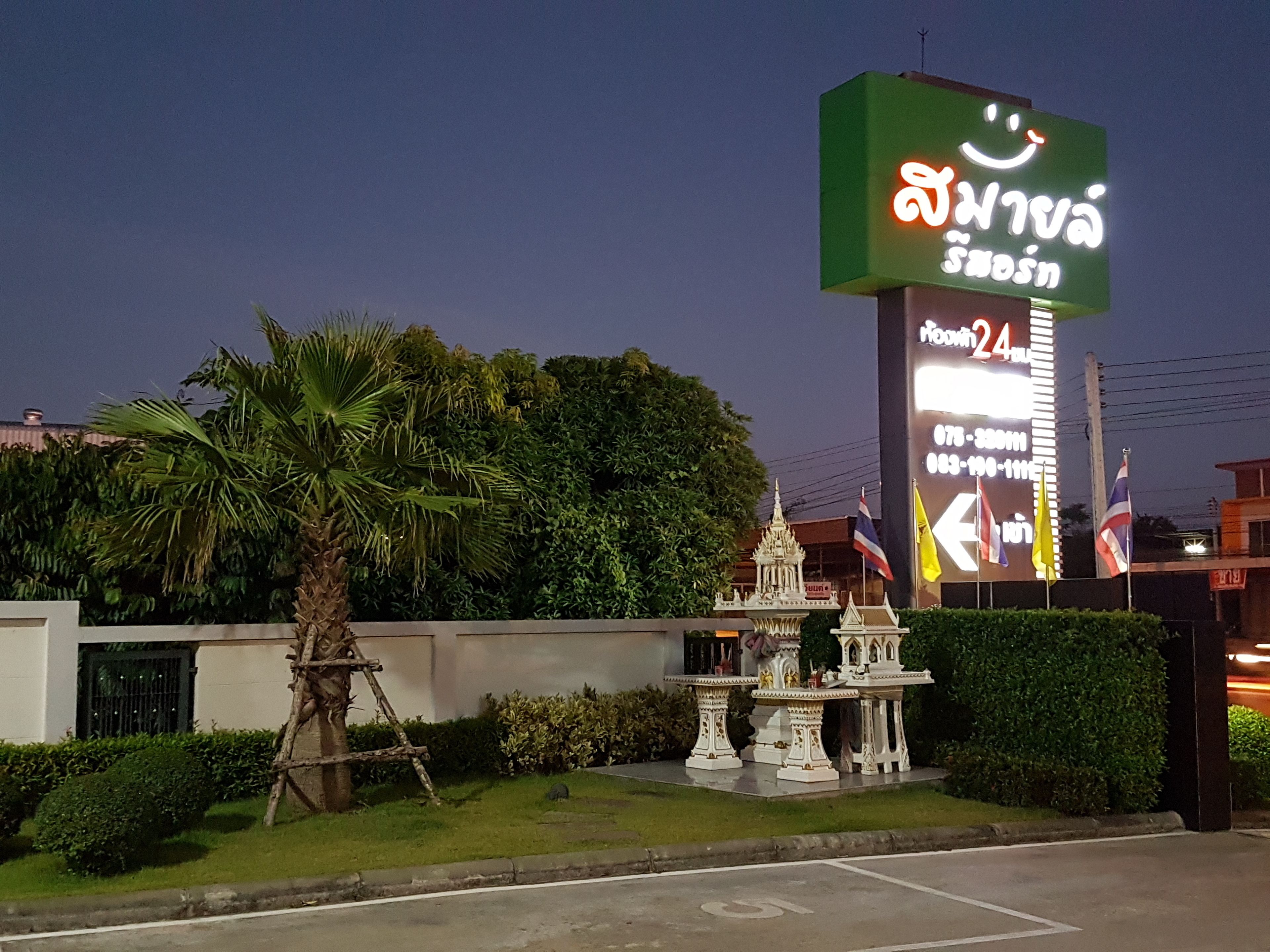 Une enseigne d’hôtel avec un visage souriant et du texte en thaï, une petite fontaine décorative et une végétation luxuriante.