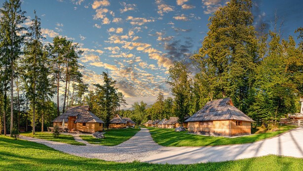 A row of rustic wooden cabins with thatched roofs, surrounded by lush greenery and tall trees, under a partly cloudy sky. - Slovenia Eco Resort (Kamnik)