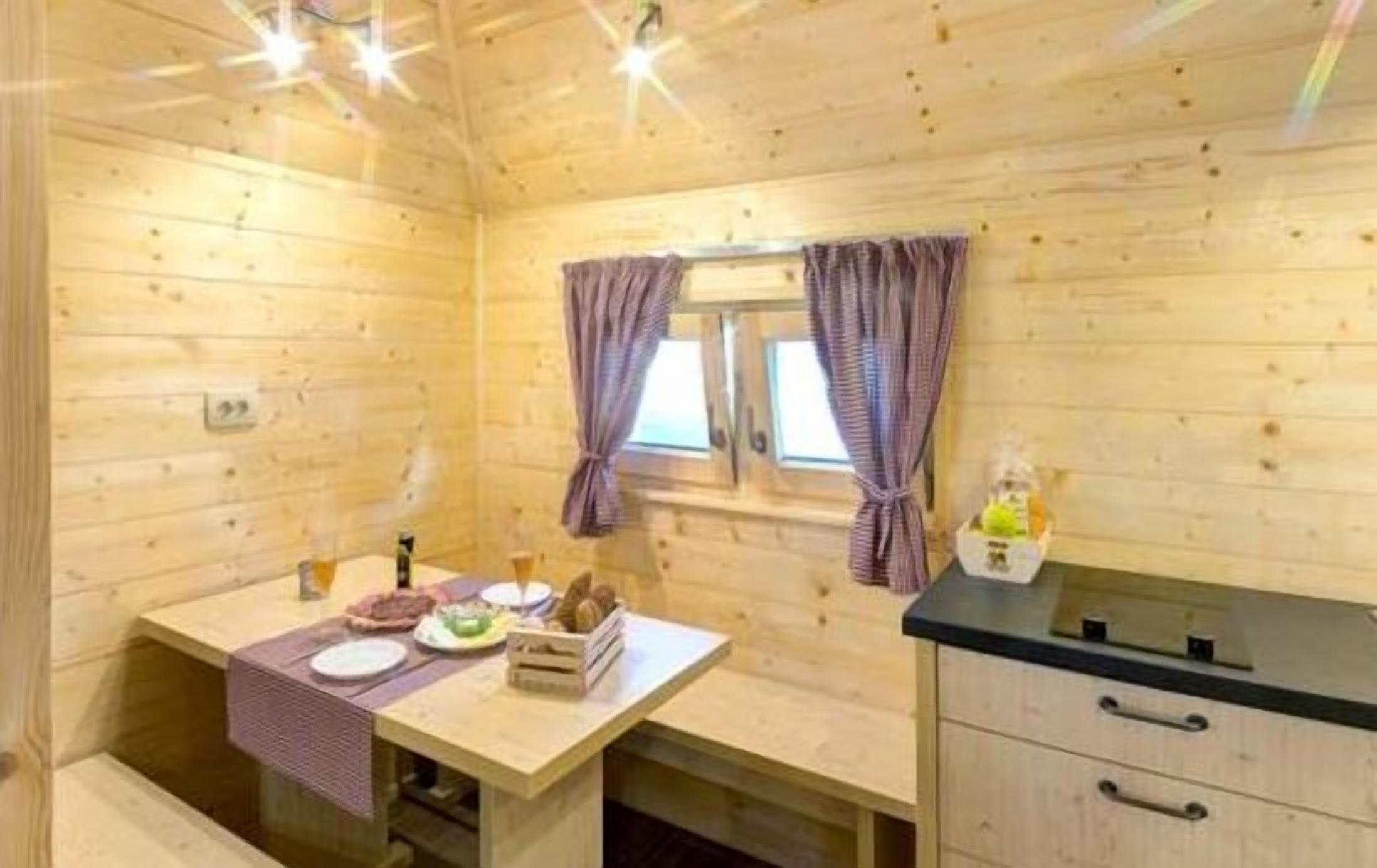 Interior de una cabaña de madera con una mesa de comedor, una ventana pequeña con cortinas y una zona de kitchenette.