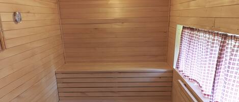 Eine Holzsauna mit einer Bank, einem Fenster mit kariertem Vorhang und einer Deckenleuchte.