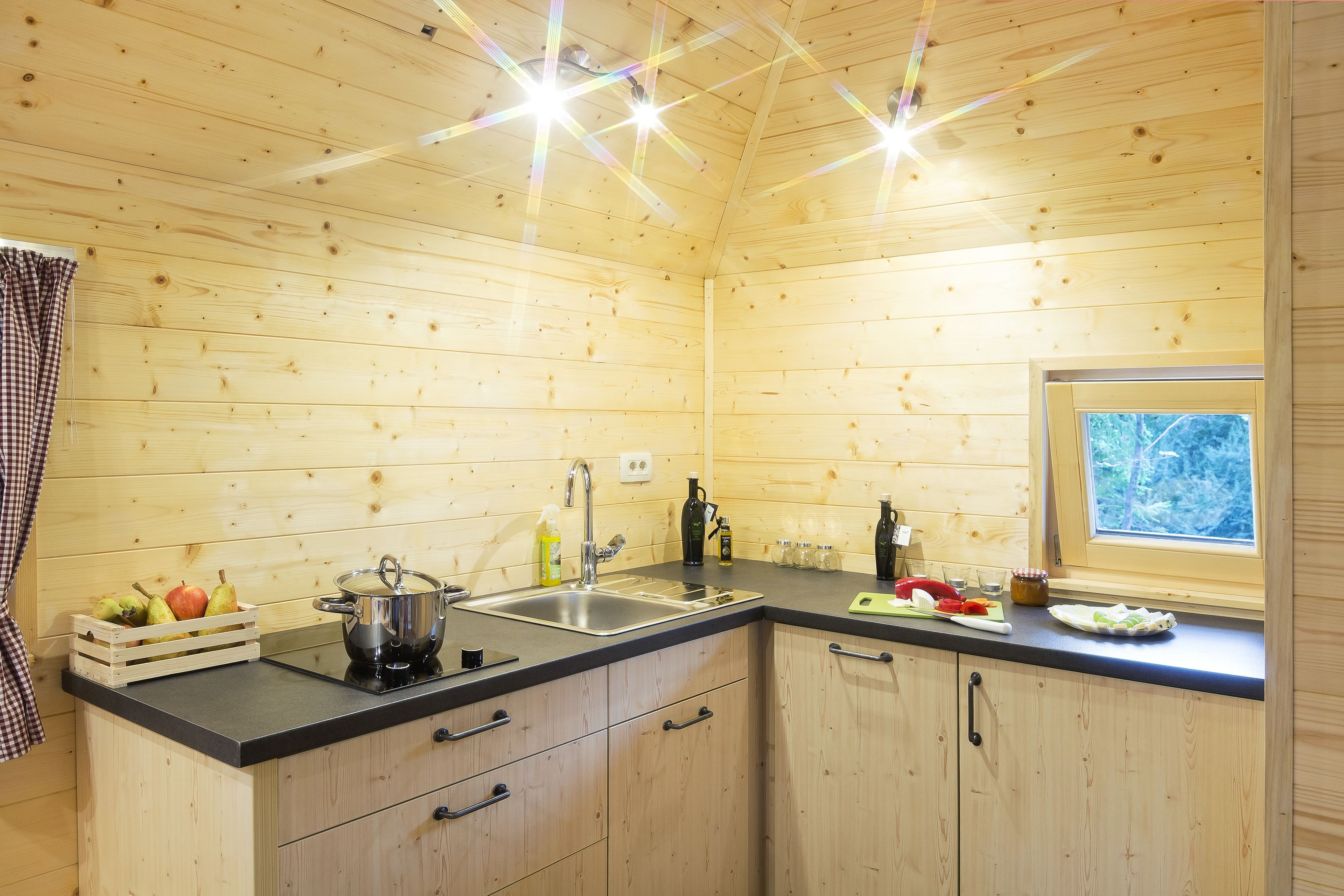 Cocina de una cabaña de madera con encimera negra, armarios de madera, un fregadero y una ventana con vistas a árboles.