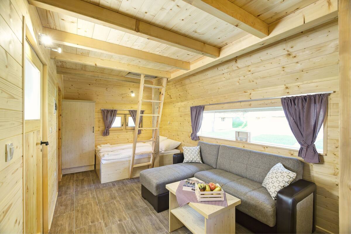 Interior de una cabaña de madera con un sofá, una pequeña mesa con fruta, una escalera y una ventana con cortinas.