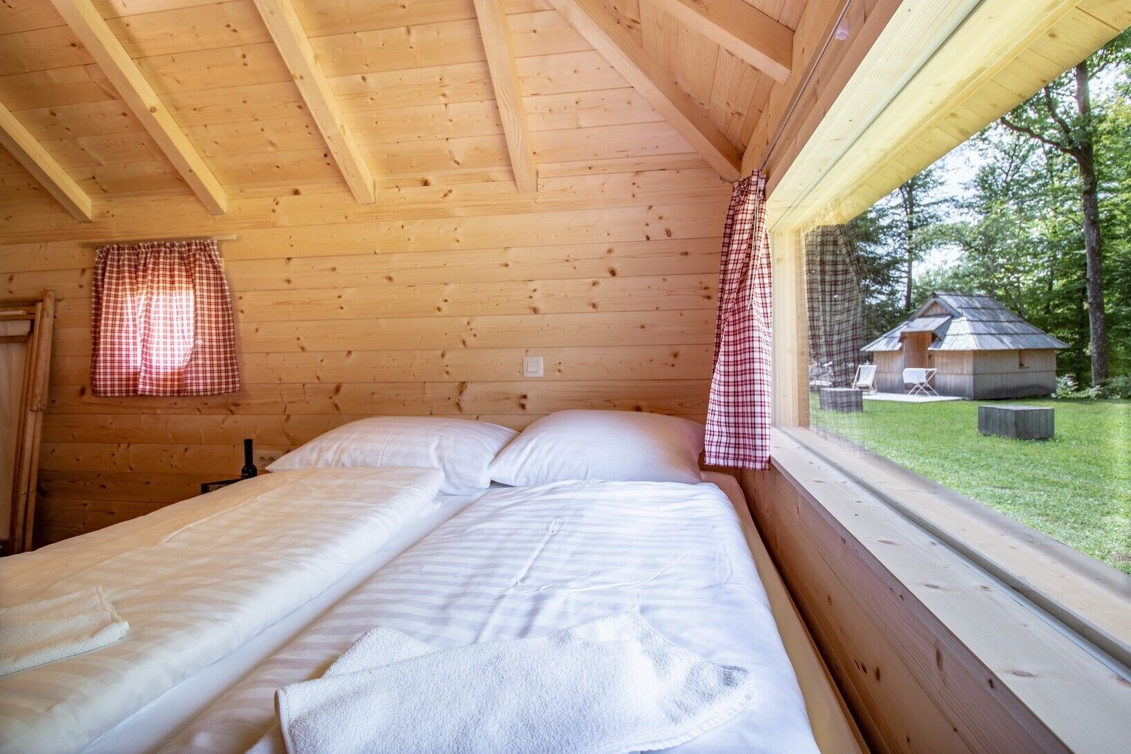 A wooden cabin room with two beds, a window with checkered curtains, and a view of a small house and trees outside.