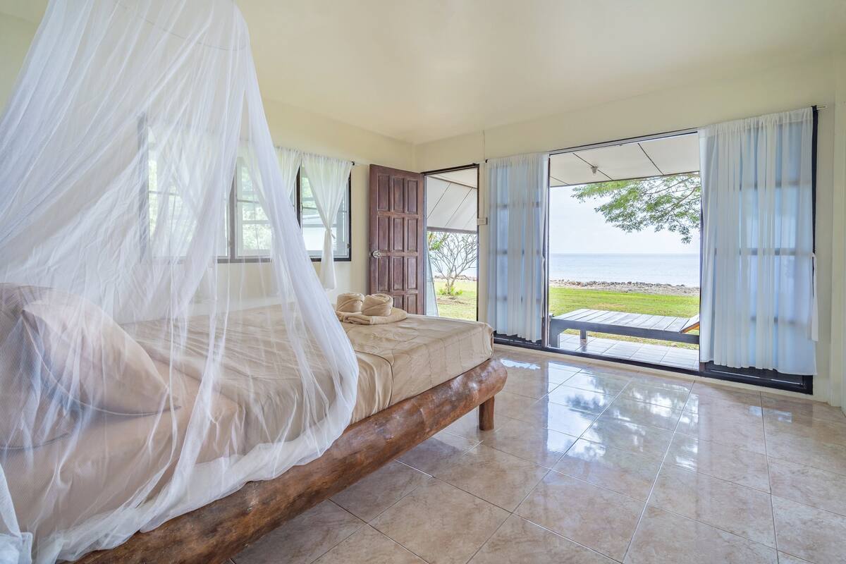 Quarto com uma cama grande, dossel e vista para o mar através de portas abertas.