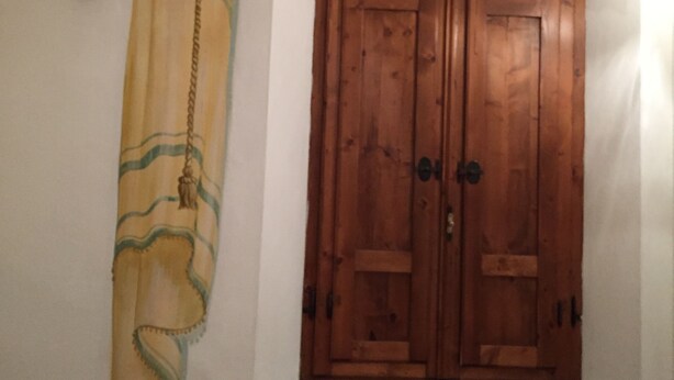 A wooden double door with a small window below it, a wall-mounted light fixture to the left, and a curtain with a tassel hanging from the ceiling. - La Locanda di Giorgia (Pisa)